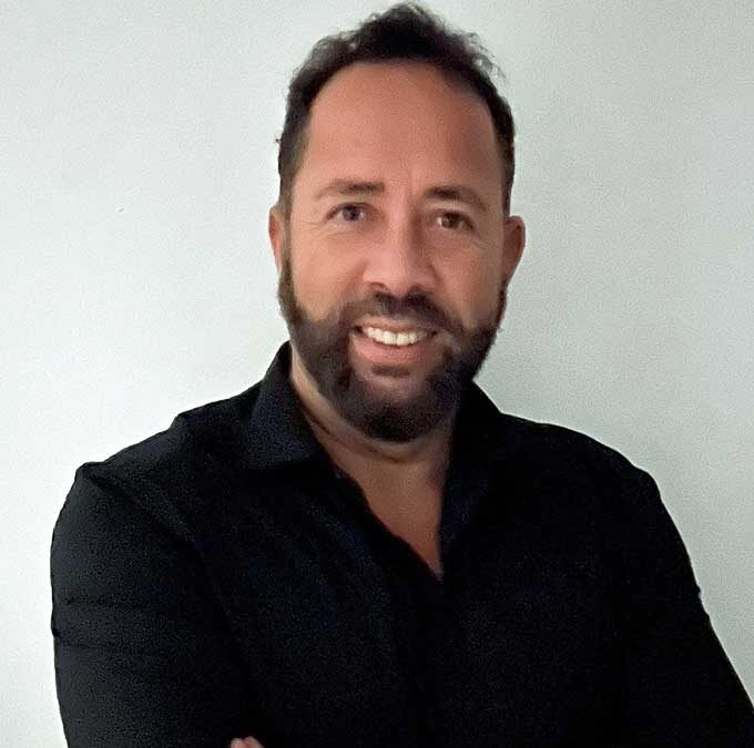 Person mit Bart trägt schwarzes Hemd und steht mit verschränkten Armen vor weißer Wand
