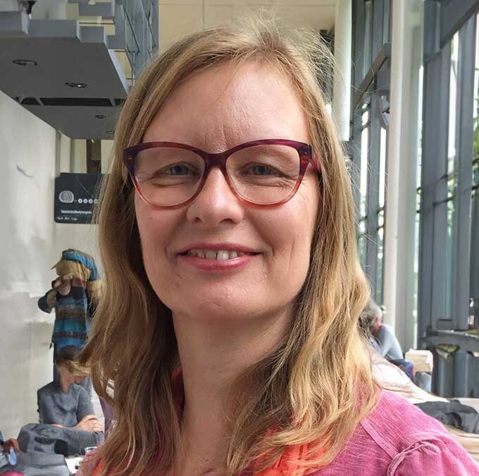 Person mit schulterlangem Haar und roter Brille trägt pinkes Oberteil, steht in einem hellen Innenraum mit großen Fenstern