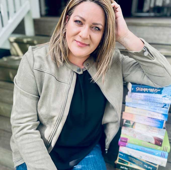 Person mit schulterlangem Haar trägt helle Jacke und Jeans, sitzt auf einer Treppe neben einem Stapel Bücher und lächelt in die Kamera