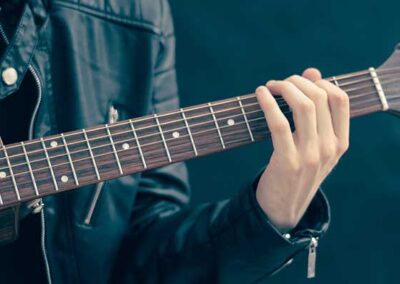 Person in schwarzer Lederjacke greift Akkorde auf Akustikgitarre, Nahaufnahme von Händen und Griffbrett