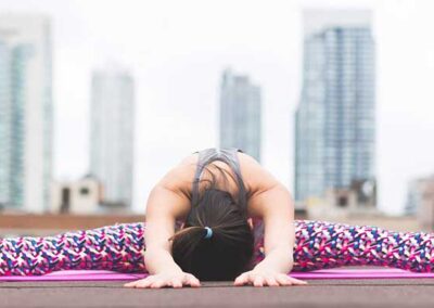 Person sitzt in weiter Grätsche und beugt Oberkörper nach vorne auf Yogamatte auf Dachterrasse mit Hochhäusern im Hintergrund